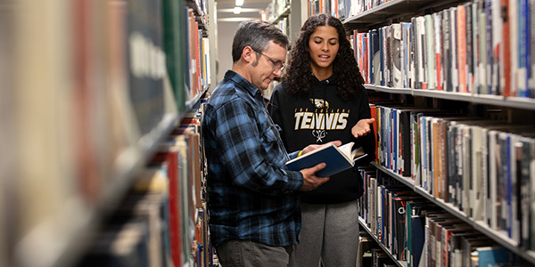 Student and professor looking at a book in library stacks